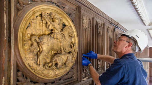A large gold coloured circular feature with a carving of a knight on a horse is being removed from a timber panelled hall by a man with a hard hat on